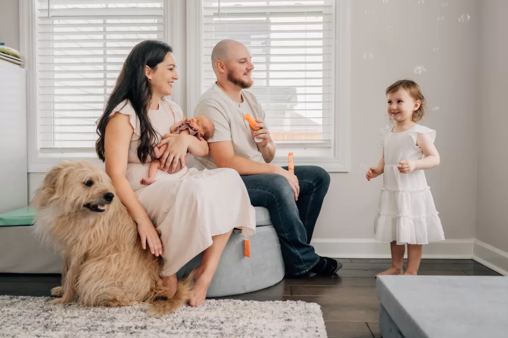 Parents holding a newborn baby looking at a toddler being silly during a newborn session. Mom is petting a big dog.