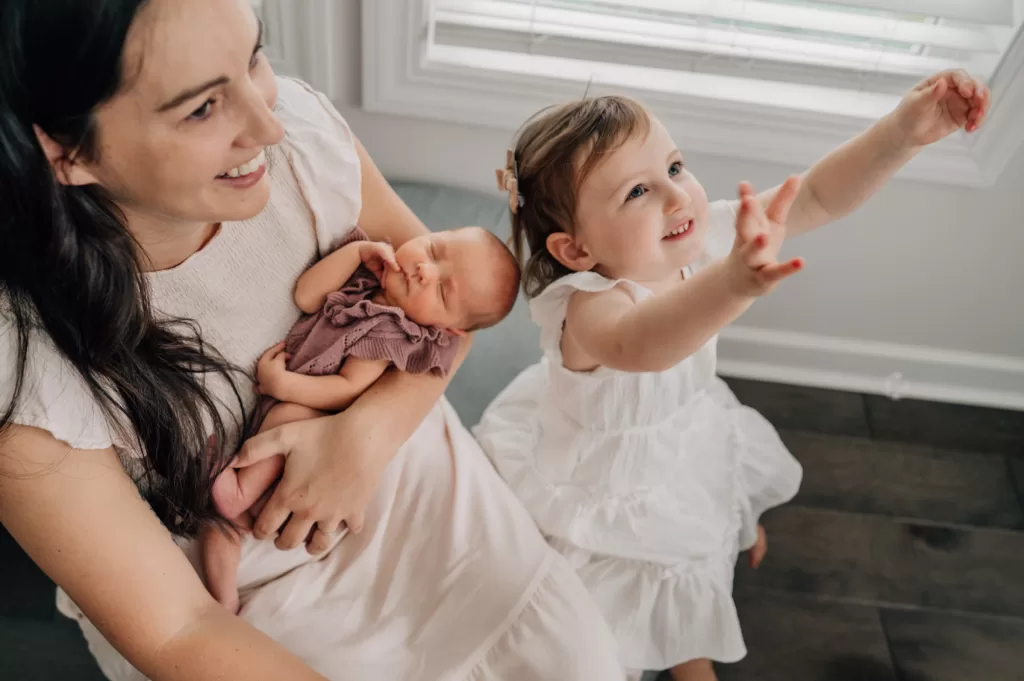 Mom smiling and holding a newborn baby girl, toddler girl reaching her hands up to catch bubbles.