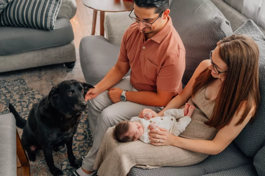 Parents sitting on a couch looking down at their newborn baby girl and a black dog.