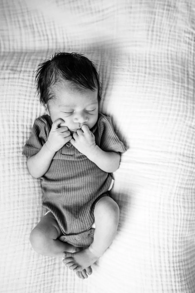 Black and white picture of a newborn baby boy laying on a white blanket with his hands close to his mouth