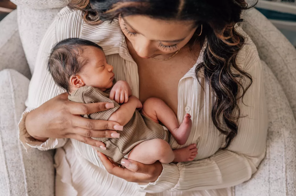 Mother holding her newborn baby boy while sitting in a beige rocking chair during a in-home newborn session