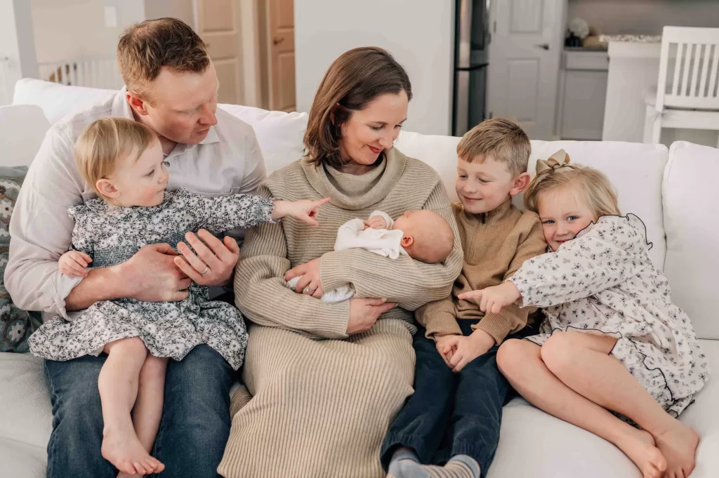 Parents and 4 children sitting on the couch together. Mom holding baby. Everyone looking down at the newborn.