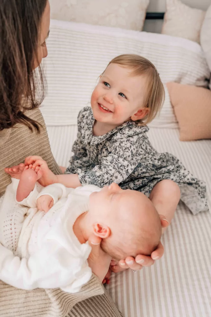 Mother smiling and looking at her toddler while holding an infant in her hands.