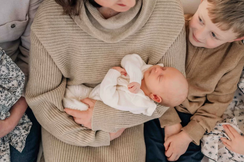 Mother holding a newborn baby, her son looking sideways at them. Planning for postpartum and the needed essentials.