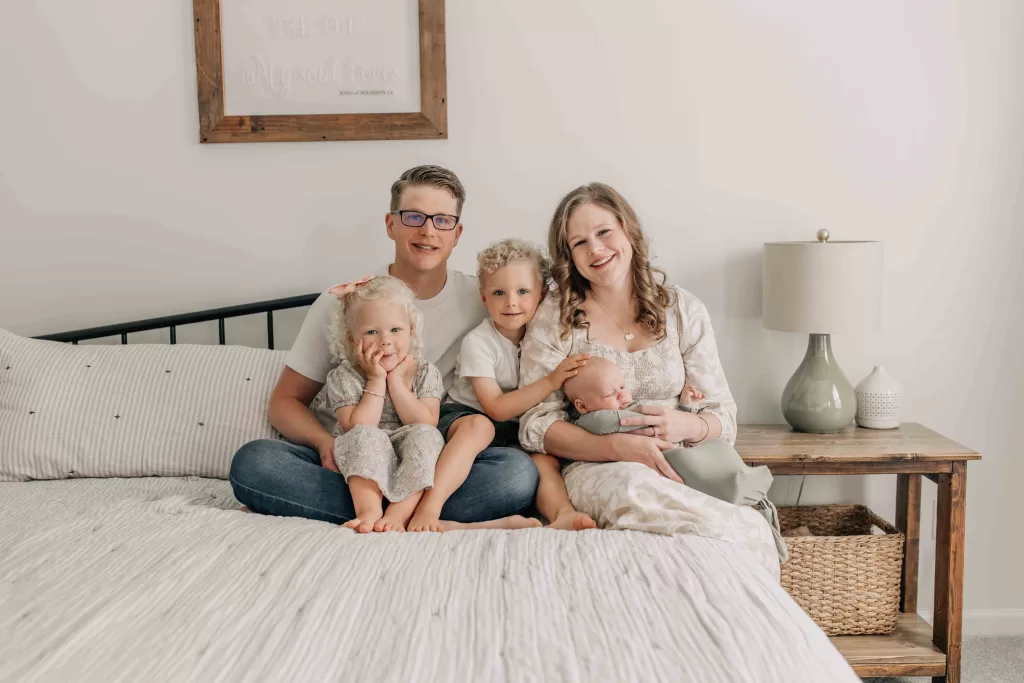 A mom and dad sit on a bed with their two toddlers and newborn in their laps after visiting baby boutiques in Columbia, SC