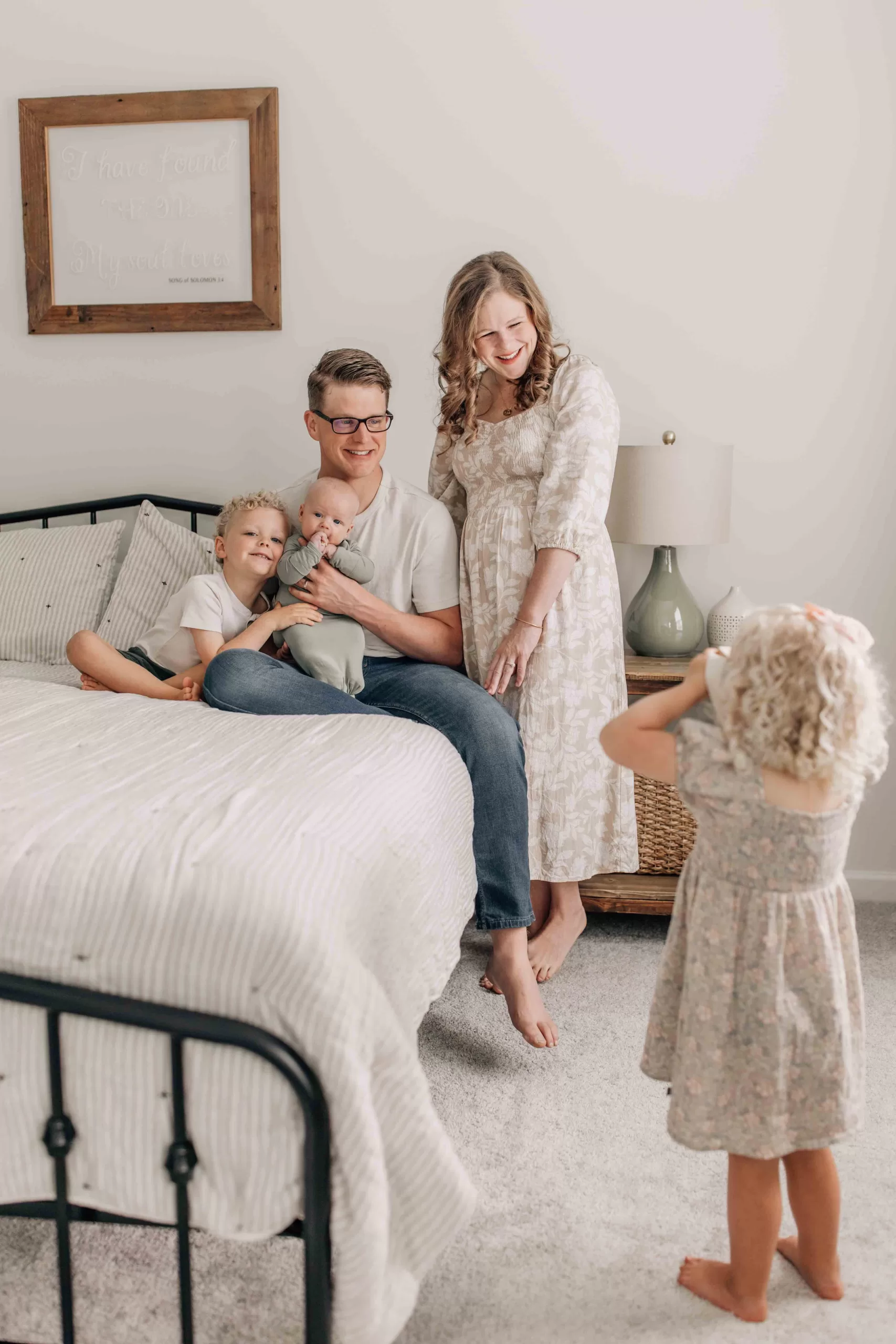 A toddler girl shows off to mom and dad snuggling on a bed with her younger brother and infant brother