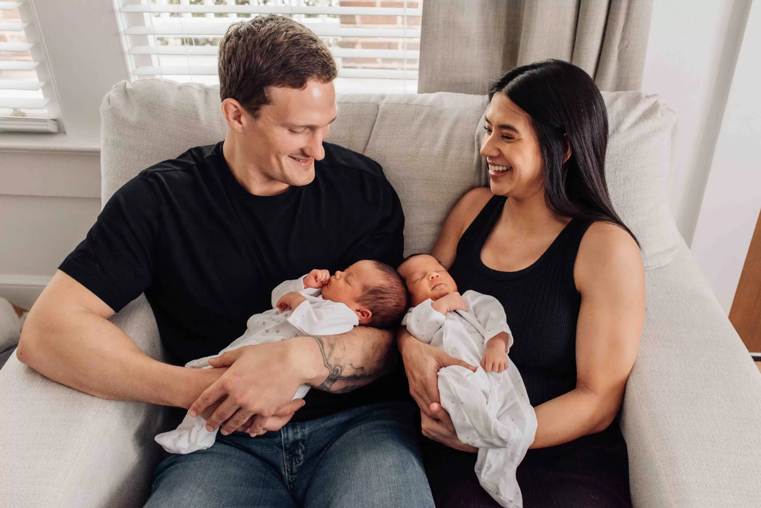 Newborn twin girls sleep in mom and dad's arms while sitting on a love seat together in black shirts after visiting pediatricians in Columbia, SC