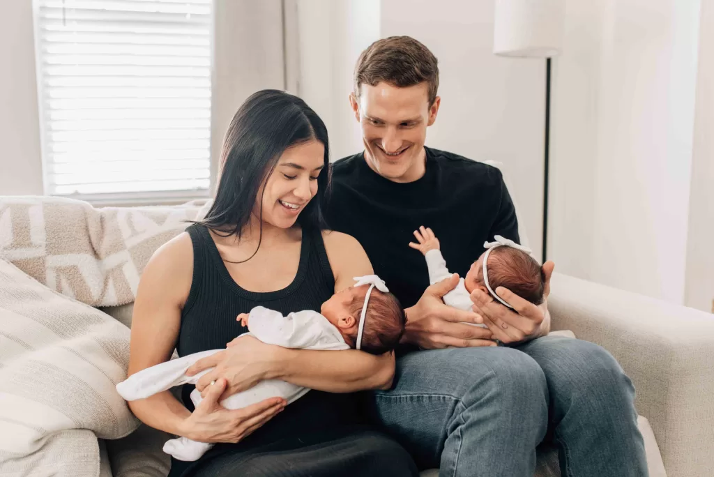 Happy mom and dad in black shirts sit on a couch holding their twin daughters in their laps after visiting pediatricians in Columbia, SC