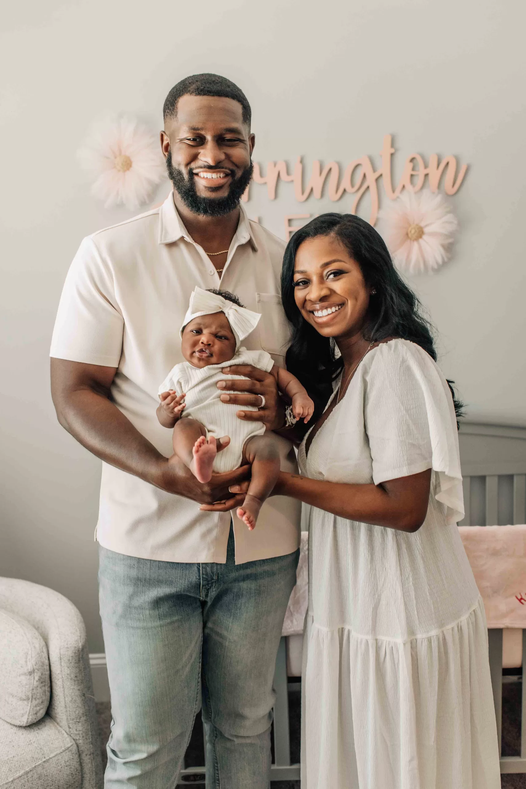 Happy new mom and dad in white stand in a nursery holding up their awake infant daughter in a white bow