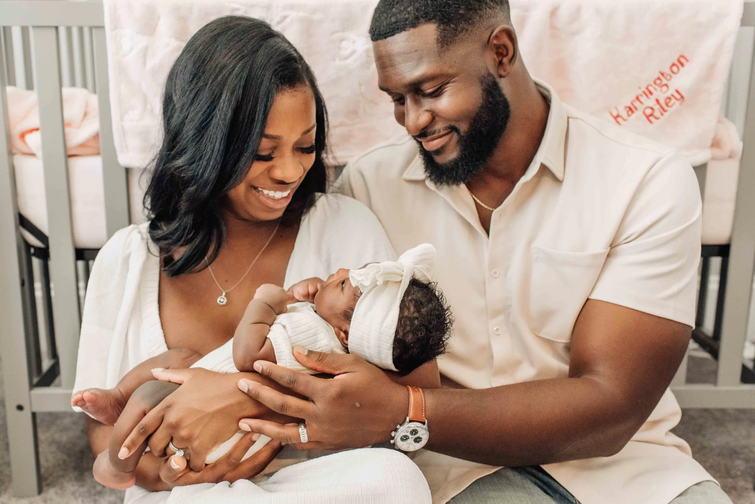 Smiling mom and dad sit on the floor cradling their infant in mom's arms against the crib after meeting postpartum doulas in columbia sc
