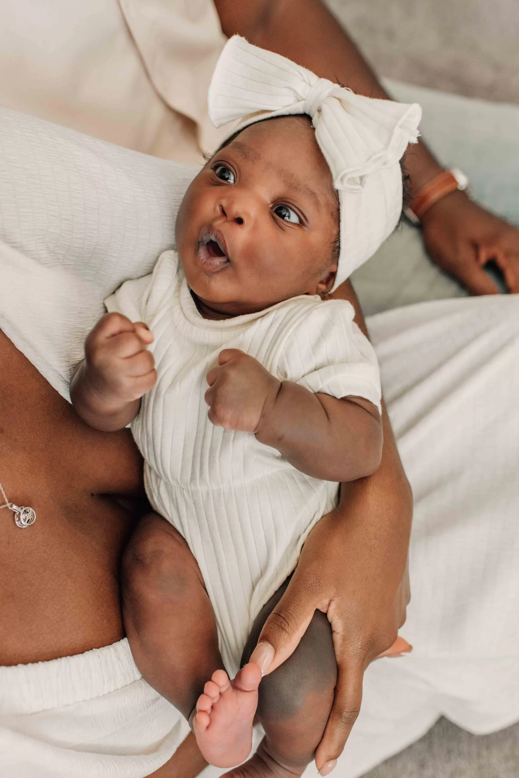 An excited infant in a white onesie and large bow lays in mom's lap on a couch next to dad after meeting postpartum doulas in columbia sc