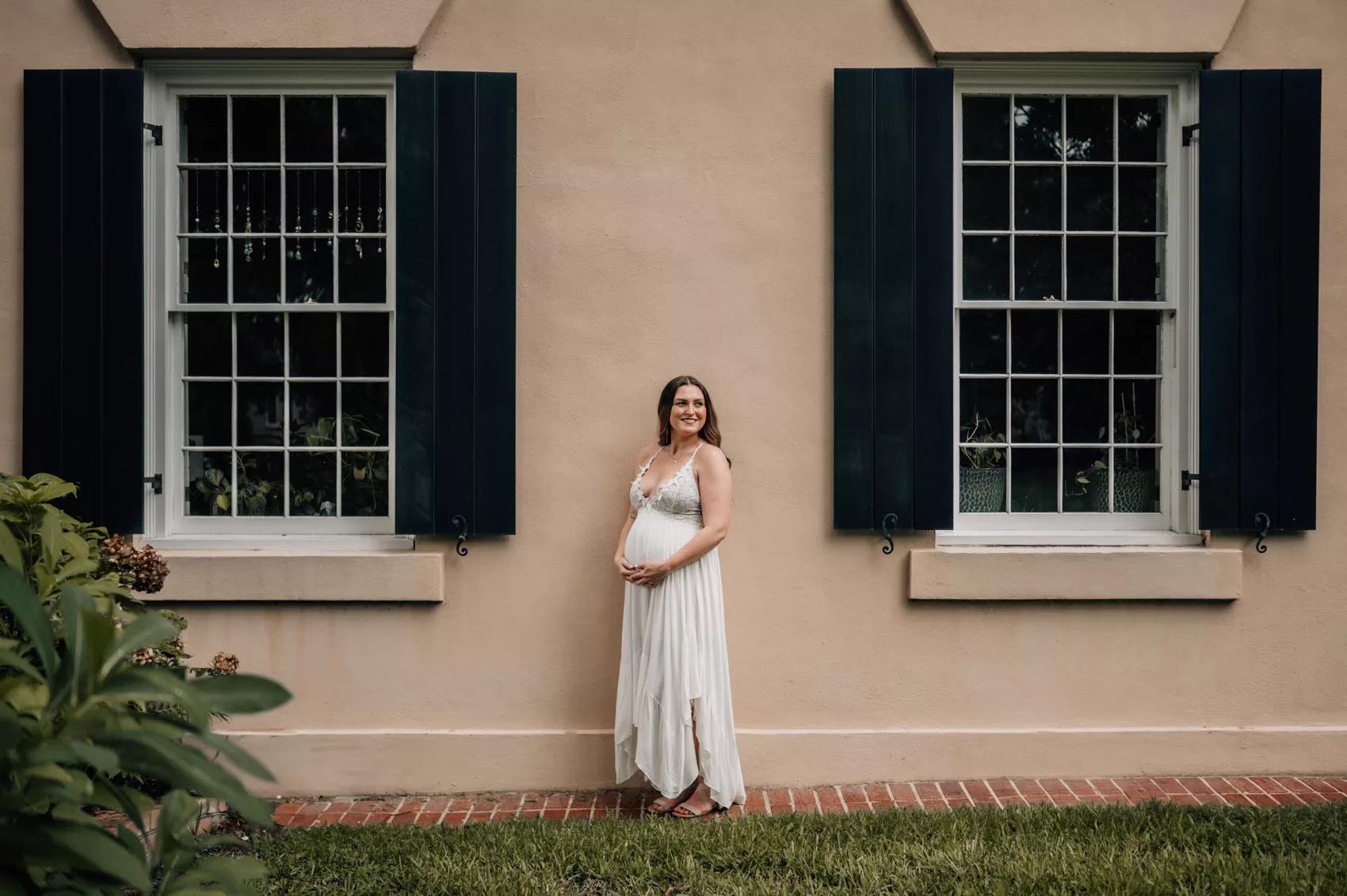 A mother to be in a white maternity gown stands between two windows of a cream building smiling over her shoulder after exploring baby shower venues in columbia, sc