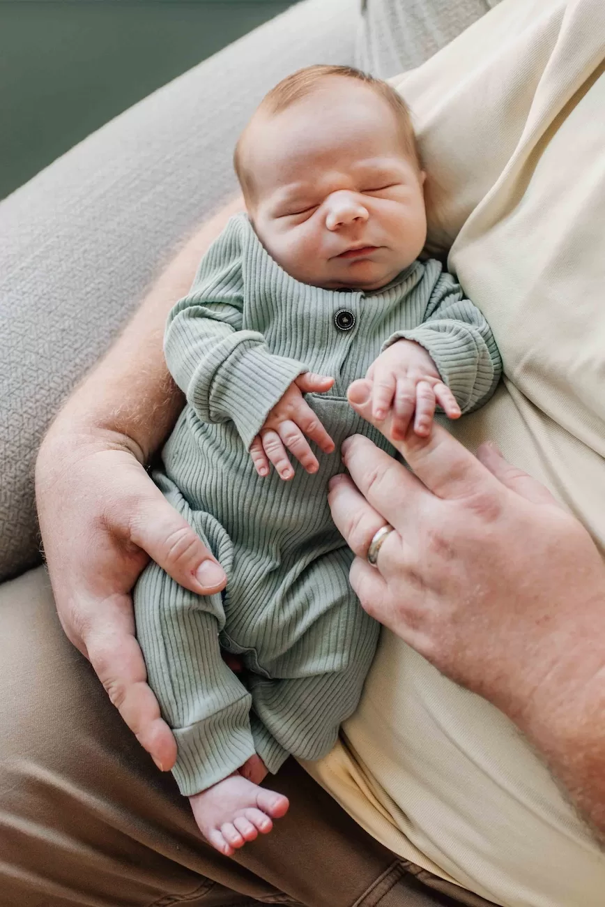 A newborn baby in a green onesie sleeps in dad's lap and holds his finger in a nursing chair after enjoying cloth diapers in columbia, sc