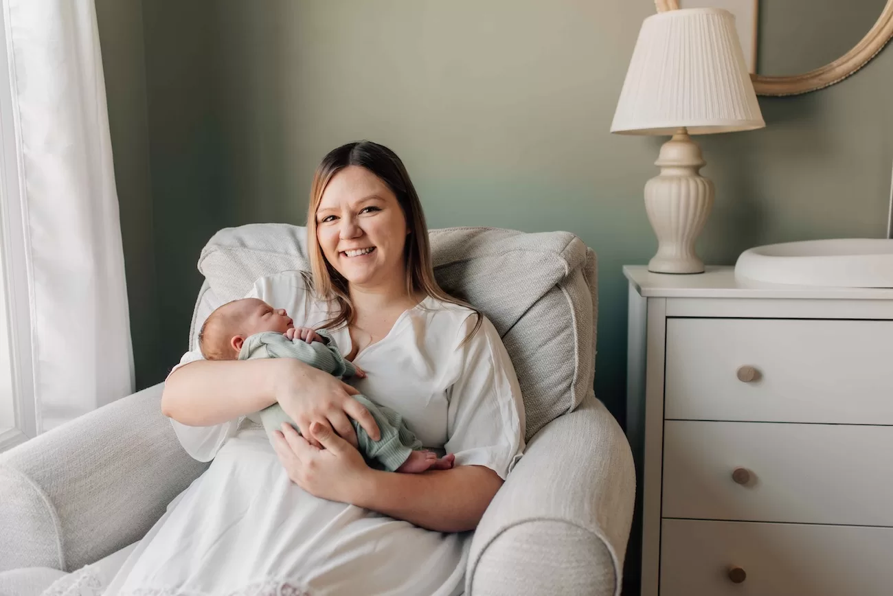 A smiling new mom in a white dress sits in a nursing chair cradling her sleeping newborn after finding cloth diapers in columbia, sc
