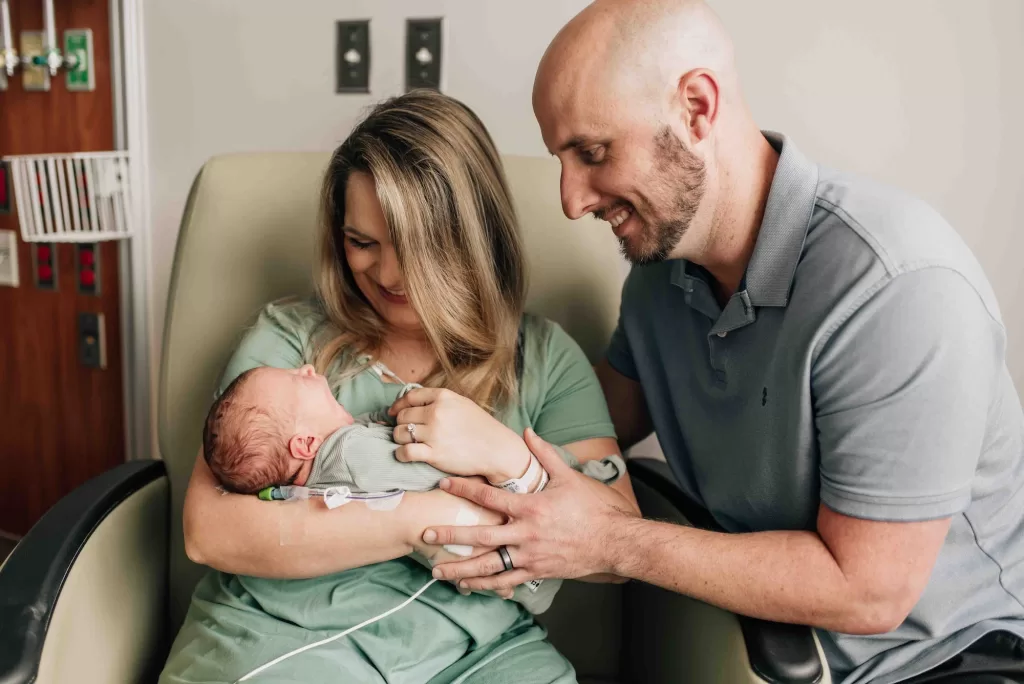 Happy mom and dad in green and blue smile down at their sleepy and yawning newborn in mom's arms in the hospital