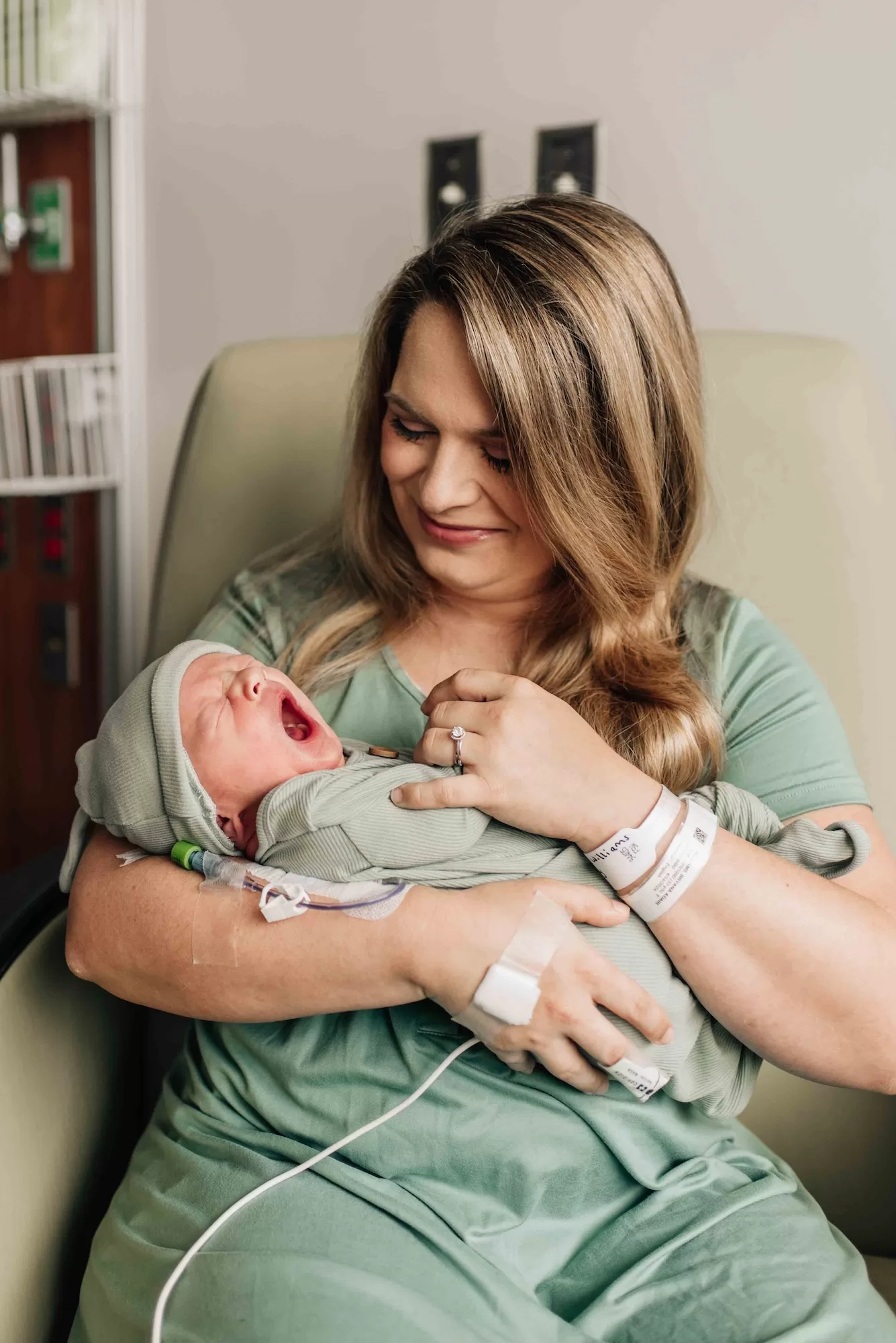 A new mom in a green dress smiles as her newborn yawns in her arms in a nursing chair thanks to doulas in Columbia, SC