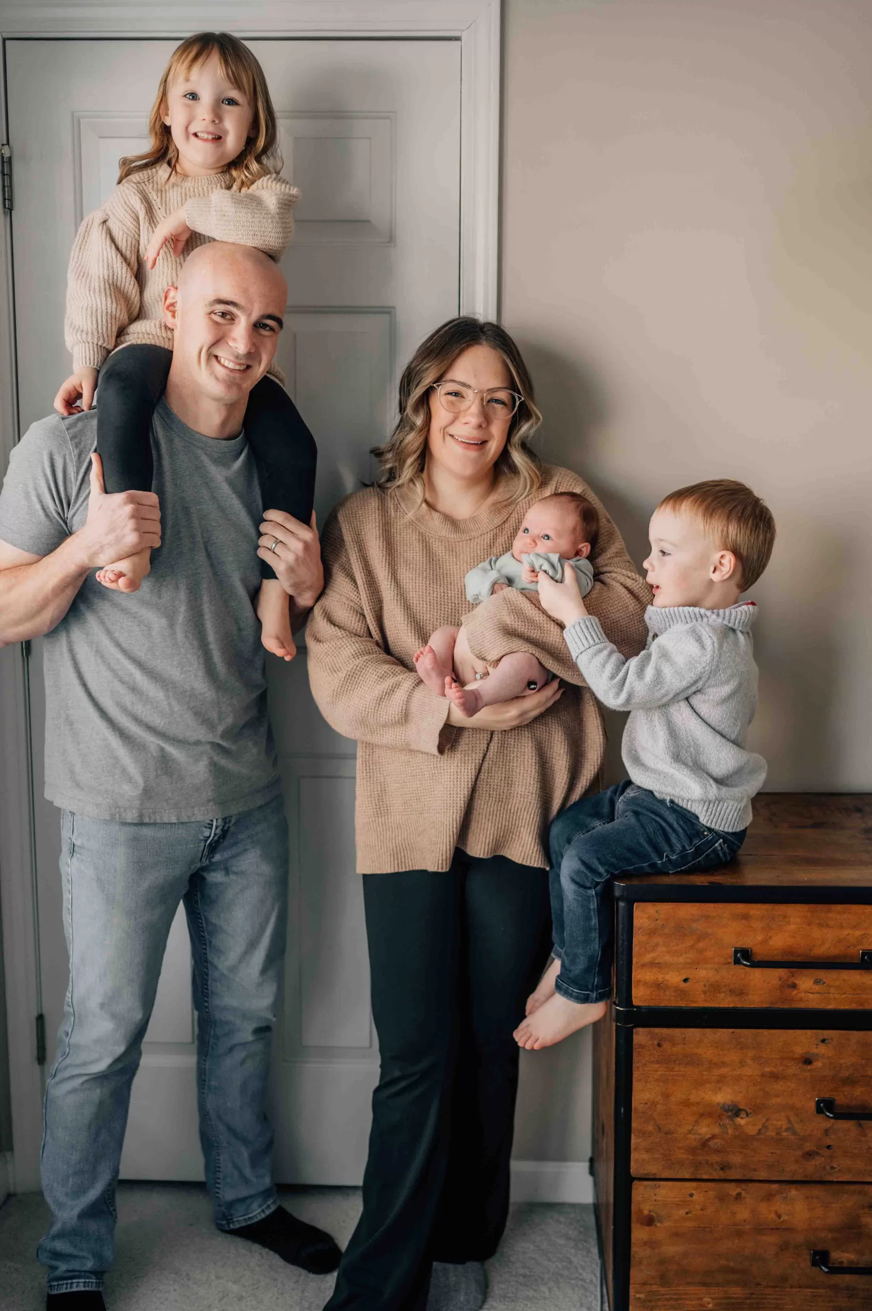 Happy mom and dad stand in their bedroom holding a newborn, a toddler on dad's shoulders and another toddler sitting on a dresser