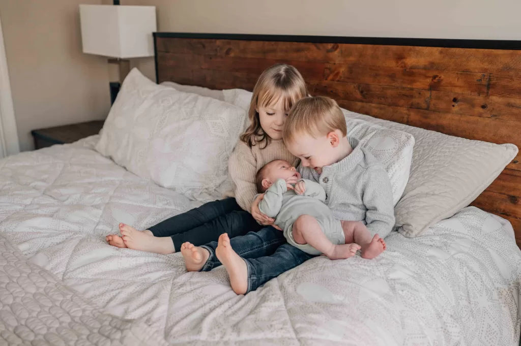 Toddler brother and sister sit on a bed smiling down to their newborn baby sibling in their laps thanks to midwives in Columbia, SC