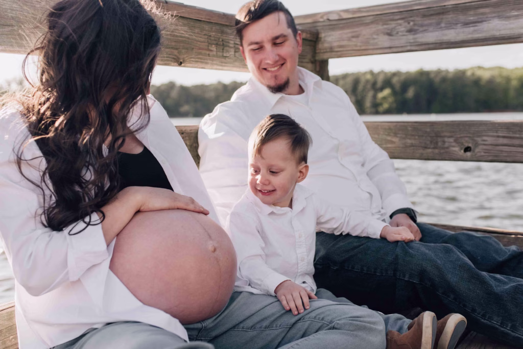 A toddler boy sits with mom and dad on a lake dock in a park smiling at mom's pregnant bump
