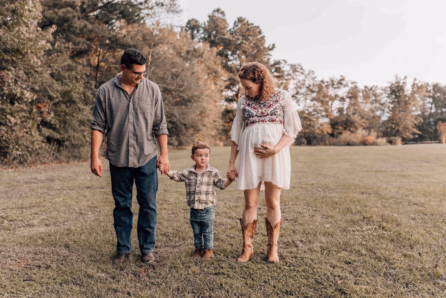 A pregnant mother walks in a park field at sunset holding hands with her toddler son in plaid and dad in jeans