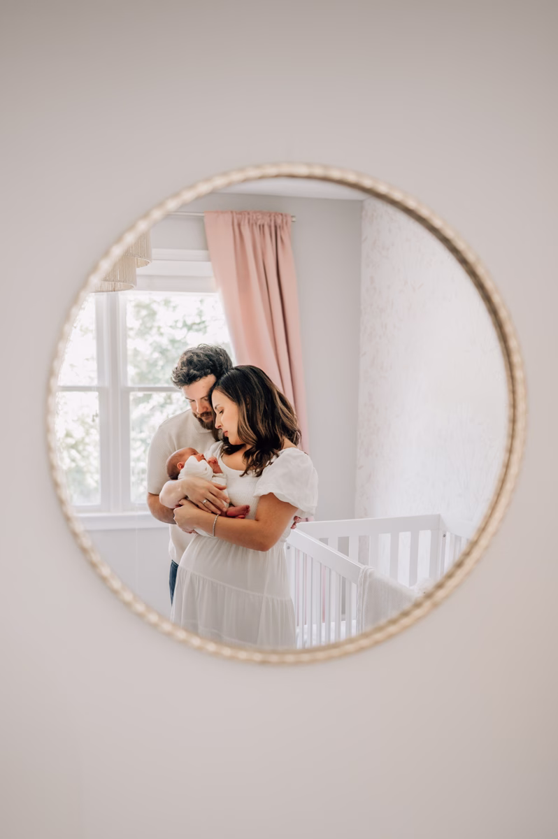 A look into a mirror at new mom and dad in a nursery cradling their sleeping newborn baby girl