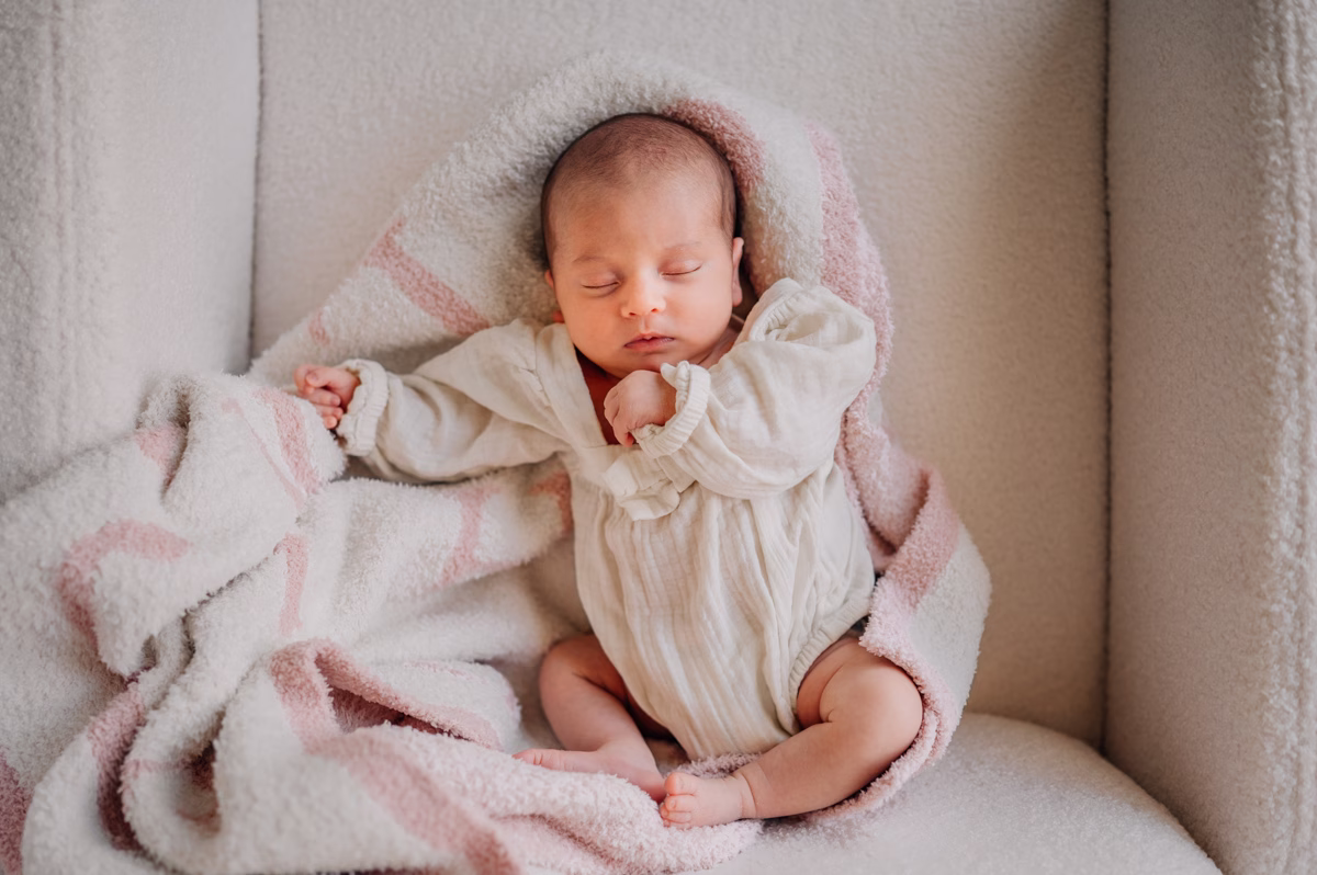 A newborn baby sleeps in a white long sleeve onesie on a pink blanket in a chair thanks to postpartum doulas in fort mill, SC
